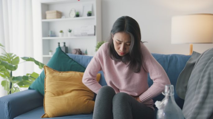 生病的亚洲女人坐在家里客厅的沙发上肚子疼。