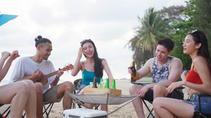 一群亚洲年轻男女在海滩上聚会。迷人的快乐朋友旅行者在海边露营时唱歌跳舞，享受热带海岛的假日度假之旅