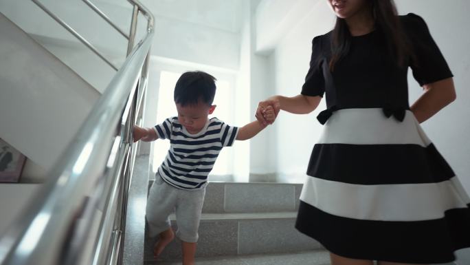 母亲牵着蹒跚学步的男孩的手走在楼梯上