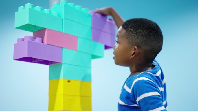 非洲黑人儿童的后视图，一名7岁男孩试图连接并添加彩色塑料积木作为机器人，没想到一些塑料积木掉了下来。