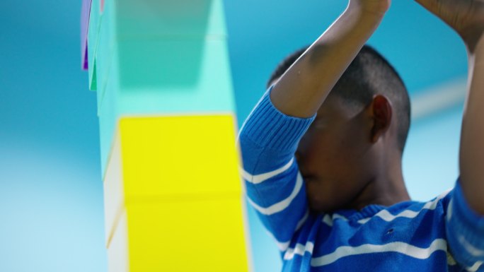 非洲黑人儿童的低角度镜头，一个7岁的男孩专注于彩色塑料积木的连接，高达头顶，并保护它在客厅里不会倒塌