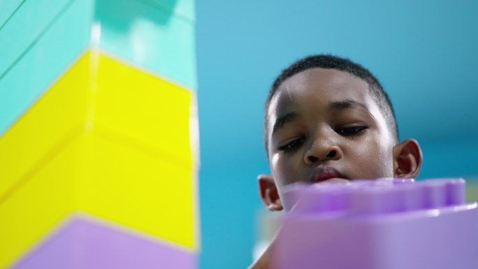 非洲黑人儿童的仰角，一个7岁的男孩打算连接并添加彩色塑料积木作为机器人，并需要保护它在客厅中不会倒塌