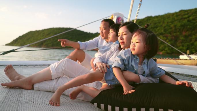 快乐的一家人享受他们的帆船旅行。他们坐在蹦床上，享受日落时的谈笑。年轻的母亲和父亲正试图向儿子们指出