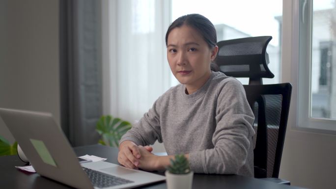 一位亚洲女性因担心问题而心烦意乱，坐在家里的办公室里看着摄像机。