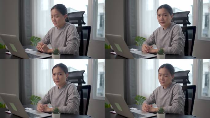 一位亚洲女性因担心问题而心烦意乱，坐在家里的办公室里看着摄像机。