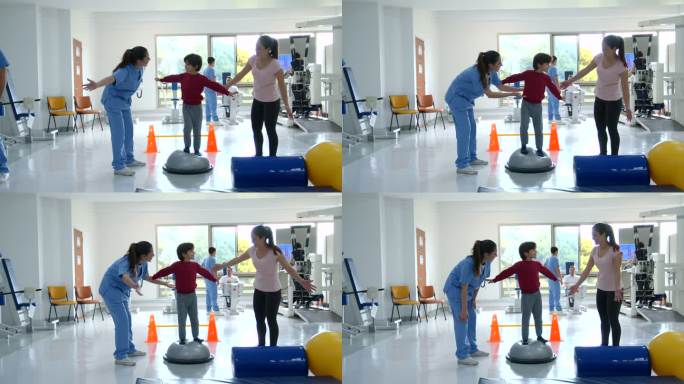 在物理治疗期间，一个可爱的小男孩按照治疗师和妈妈的指示，试图站在锻炼的半边球上