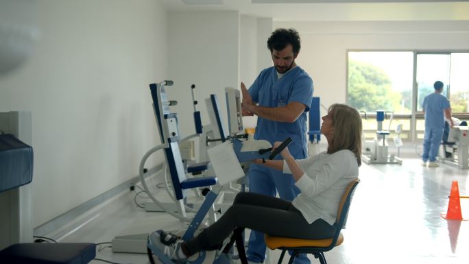 老年妇女在物理康复中心接受物理治疗时骑着静态自行车，一边听男性治疗师解释动作