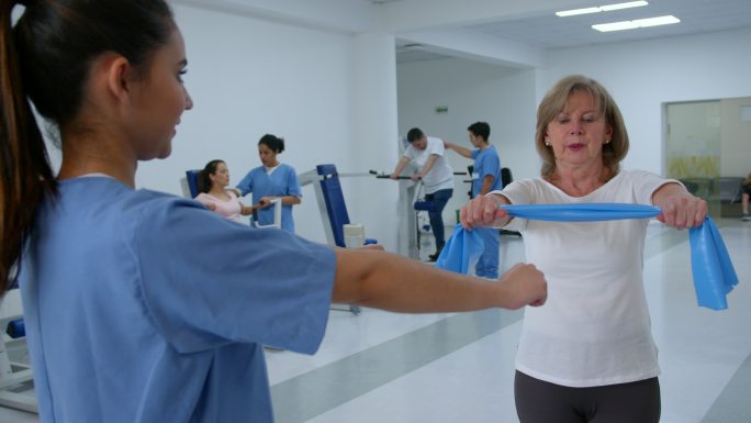 一名老年妇女在康复诊所按照治疗师的指示，用阻力带锻炼手臂