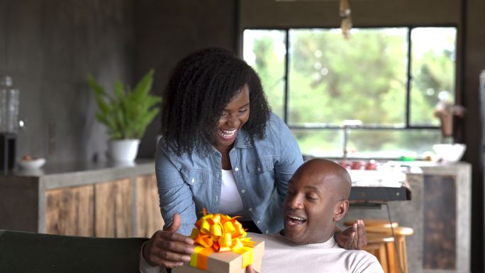 一位可爱的黑人女性在情人节给她的伴侣送礼物，让他惊喜不已，而他正在智能手机上发短信，在家里的沙发上放