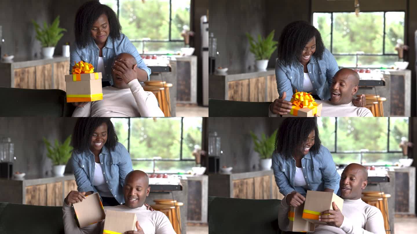 一位可爱的黑人女性在情人节给她的伴侣送礼物，让他惊喜不已，而他正在智能手机上发短信，在家里的沙发上放