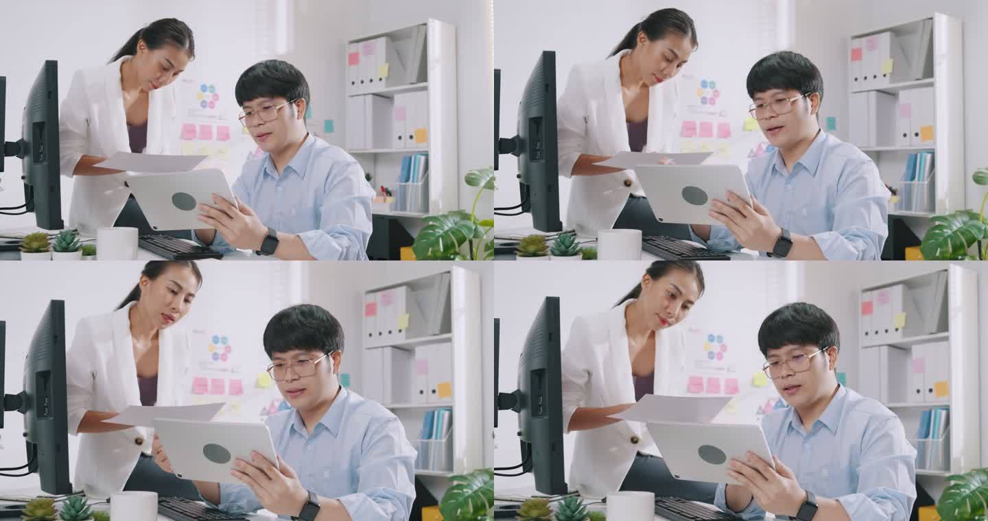 年轻的亚洲商人坐在电脑桌前，看着数字平板电脑的书写，在小工作场所为他的女同事检查工作报告、营销和演示