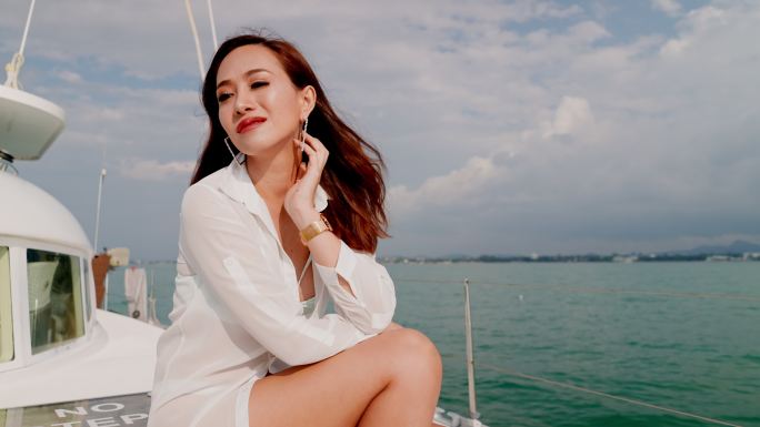 一位美女穿着白色束腰外衣在游船上放松，举手保护脸部免受阳光照射，享受美景。海上暑假。