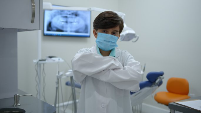 一个可爱的小男孩在牙医那里练习假装穿着实验服，戴着防护面罩，交叉双臂面对镜头