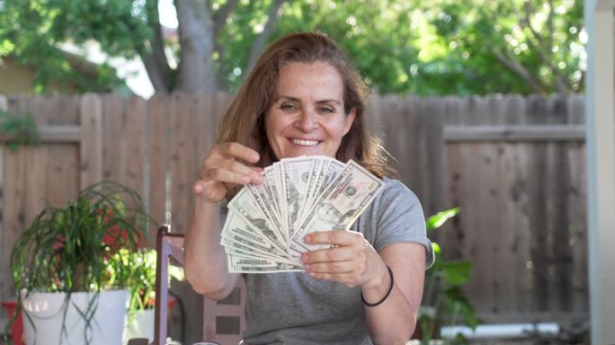 微笑的成熟女人拿着一堆美元钞票坐在后院
