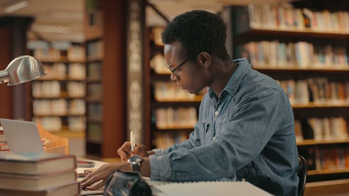 一个年轻的男学生在大学图书馆做项目研究或准备考试时做笔记。自信的法律系学生独自看书
