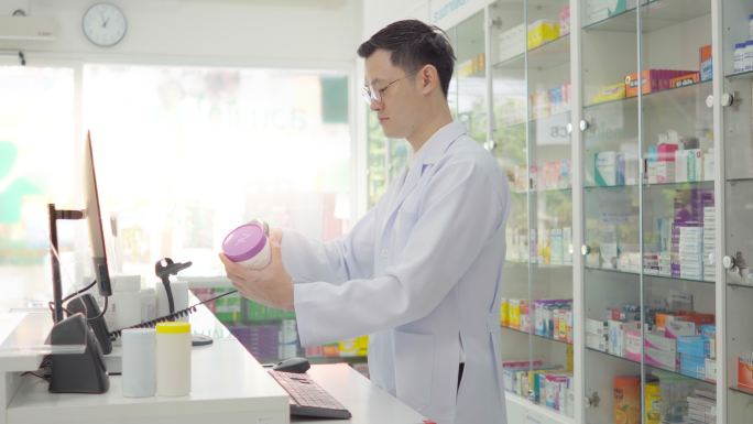 药房药房背景下的亚洲药剂师肖像。药剂师微笑着，用电脑检查药房的库存，以便销售药片，并分配给患者。医疗