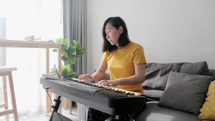 亚洲成年母亲在家中靠窗的沙发上通过手机应用程序学习键盘乐器，生活方式理念。