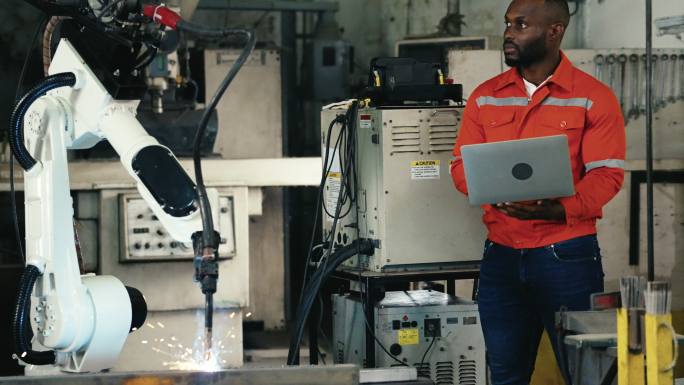 在制造业中，非洲裔美国工程师用笔记本电脑控制和编程自动化机器人手臂机器。