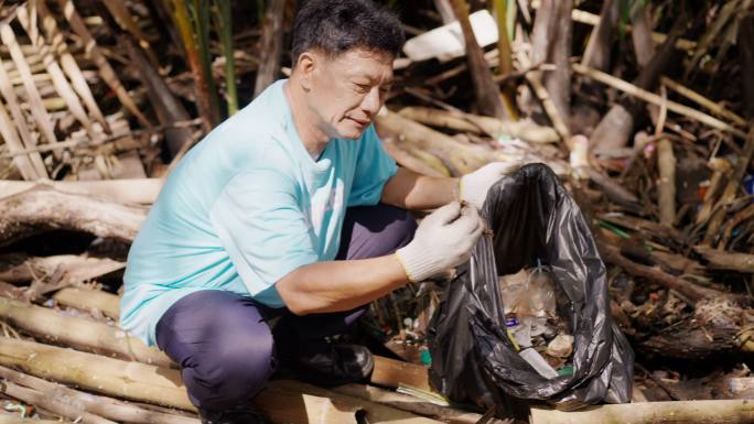 亚洲老年人志愿者在红树林中故意将垃圾和塑料袋收集到垃圾袋中，同时对人们乱扔垃圾感到非常难过，以拯救地