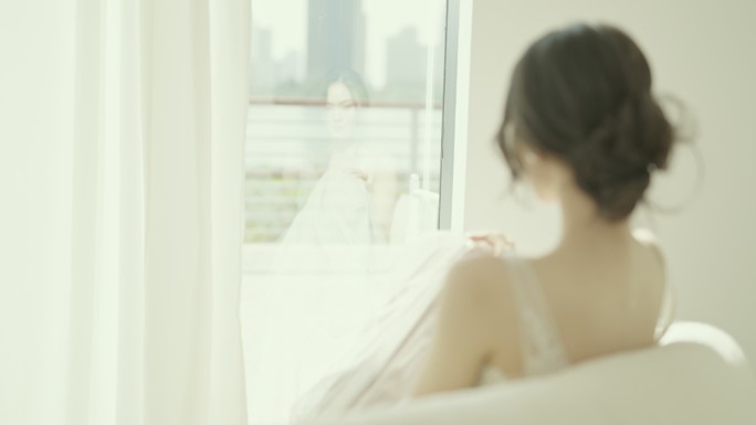 身穿白色连衣裙的美丽亚洲新娘坐在落地窗前的沙发上，慢镜头拍摄