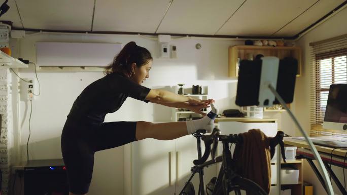 亚洲女自行车运动员，她在家锻炼。她骑自行车模拟器。她伸懒腰