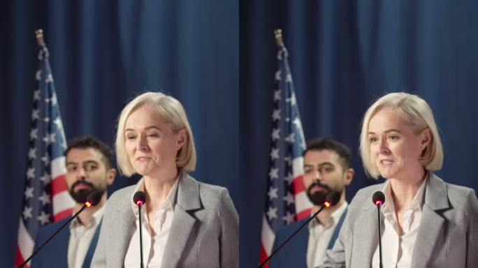 一位身穿灰色夹克的金发女政客在辩论会上发表演讲，我们看到一位秘书站在她身后