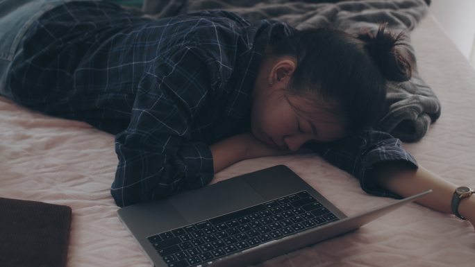 亚洲女商人在家工作，带着笔记本电脑，很累，在卧室的床上感到困倦。社会疏远的概念是采取感染控制行动，阻