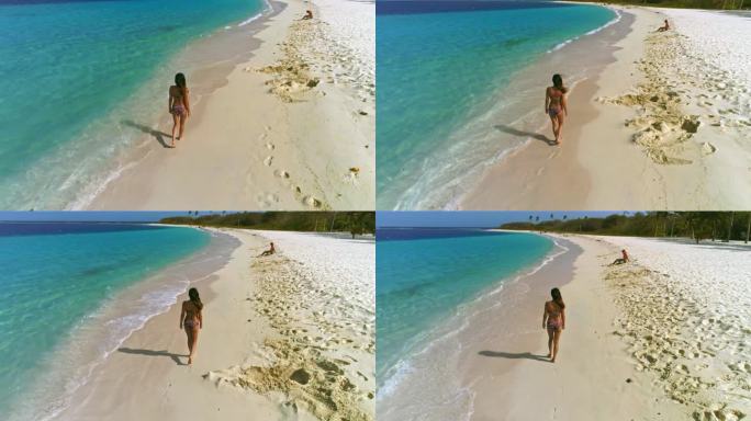 鸟瞰一位西班牙裔年轻女子在热带岛屿海滩上悠闲地散步，海滩上有椰子树、沙滩、海浪和碧绿的海水
