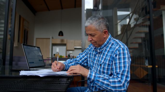 一位老人坐在家中的室外露台上，一边在笔记本电脑上查看银行对账单，一边在纸上做笔记