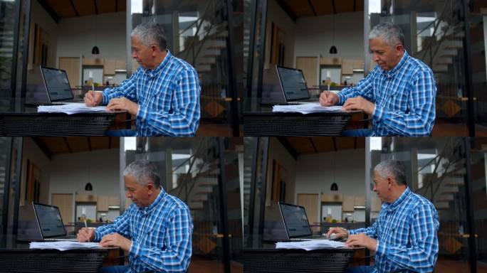 一位老人坐在家中的室外露台上，一边在笔记本电脑上查看银行对账单，一边在纸上做笔记