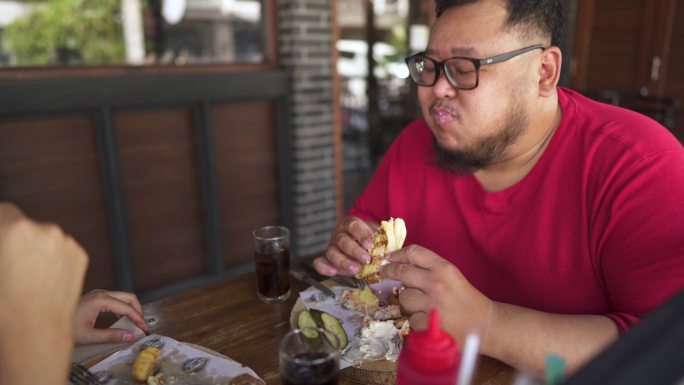 一个胖胖的年轻人在午餐时和朋友一起吃汉堡包，谈笑风生。