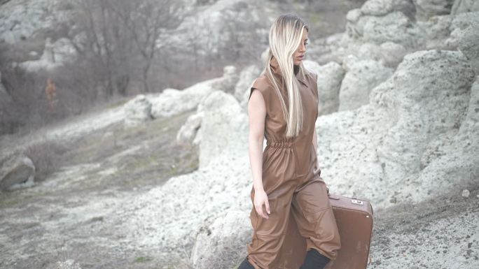 棕色连体衣的女性美女在岩石地形上携带一个匹配的手提箱