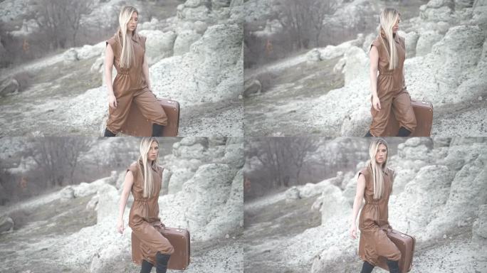 棕色连体衣的女性美女在岩石地形上携带一个匹配的手提箱