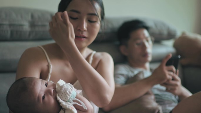 周末，亚裔中国母亲在客厅用奶瓶喂养男婴，而父亲在看他的智能手机
