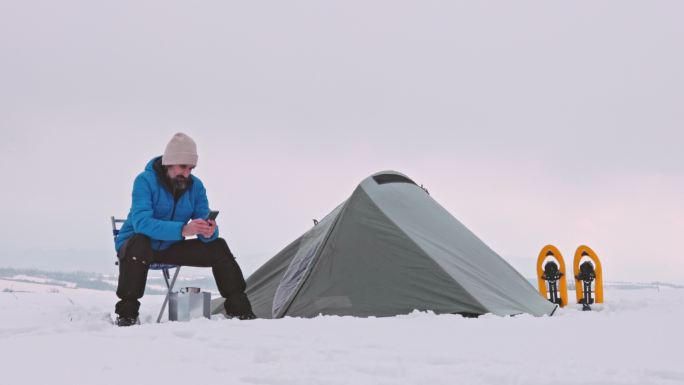 成熟的游客一边用手机喝咖啡，一边在冬天独自在大自然中的山上露营。