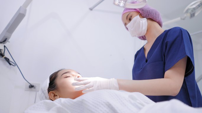 侧视图：一位医生戴着卫生手套，戴着发帽，为年轻的亚洲女性提供医疗服务，将手术灯调整到面部，检查皮肤质