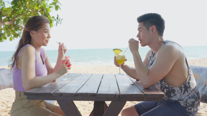 亚洲年轻男女情侣一起在海滩上喝果汁。迷人的新婚夫妇坐在海边的餐桌上，享受热带海岛的度假蜜月之旅。