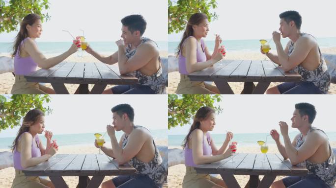 亚洲年轻男女情侣一起在海滩上喝果汁。迷人的新婚夫妇坐在海边的餐桌上，享受热带海岛的度假蜜月之旅。