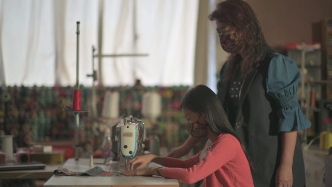 亚洲华裔高级女教师指导年轻女学生用缝纫机在录音室教育课上缝制蜡染织物防护面罩