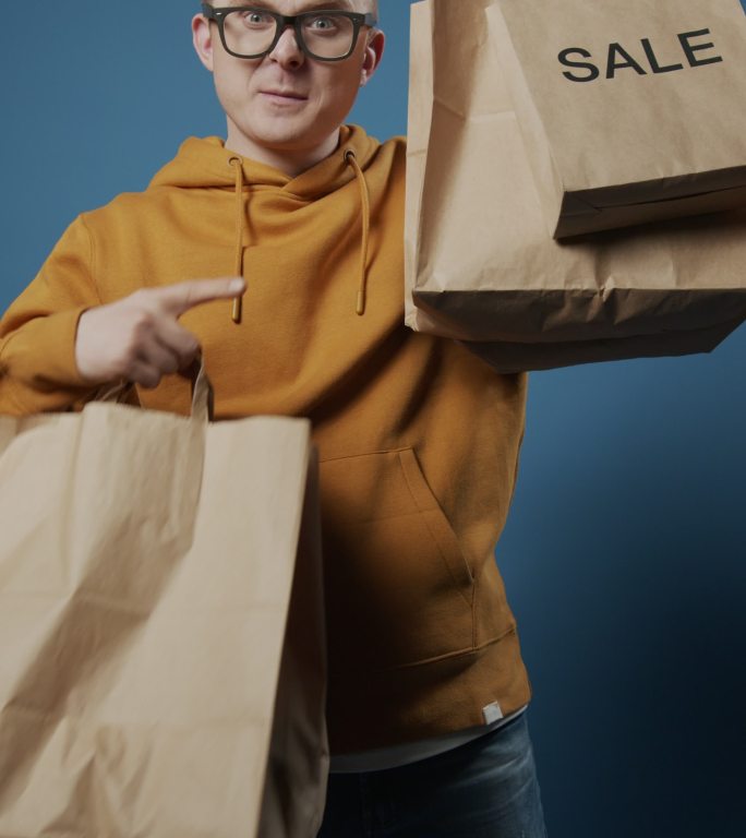一位快乐的金发流行博主站在蓝色背景下，在镜头前展示他的购物袋