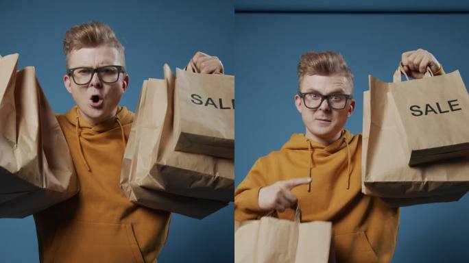 一位快乐的金发流行博主站在蓝色背景下，在镜头前展示他的购物袋
