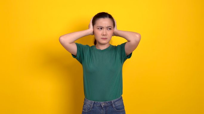 一名亚洲妇女感到厌烦和恼怒，用黄色背景遮住了自己的耳朵。