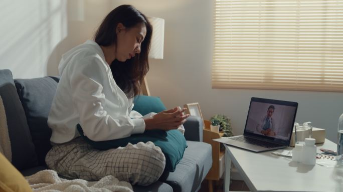生病的年轻亚洲妇女在家里的客厅里用笔记本电脑和坐在沙发上的医生交谈。