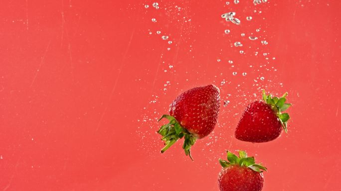 有机水果，成熟多汁的草莓在粉色背景下落入水中。美味新鲜的草莓滴入起泡饮料中。健康营养的饮食
