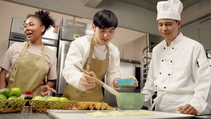 快乐的年轻成人Asain男子学生在糕点班的工作坊里尝试烹饪，在糕点面团上涂上打好的鸡蛋，而他的同学在
