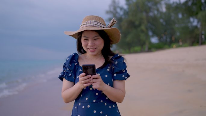 亚洲成年女性在沙滩上散步时，在智能手机上随意地微笑着在社交媒体平台上发布照片和文字，同时在沙滩上漫步