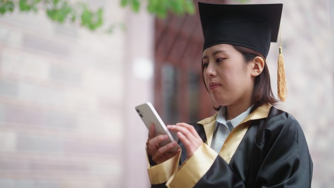 身穿毕业礼服的亚洲女大学生在校园里使用智能手机发短信