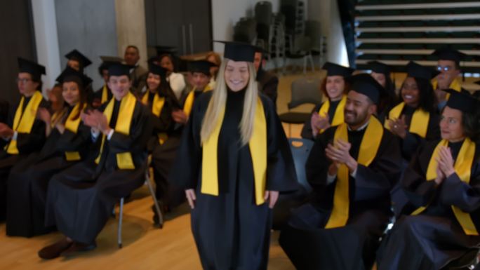 美丽的女学生从椅子上站起来准备去领取大学毕业证书，而她的同学们则非常高兴地鼓掌