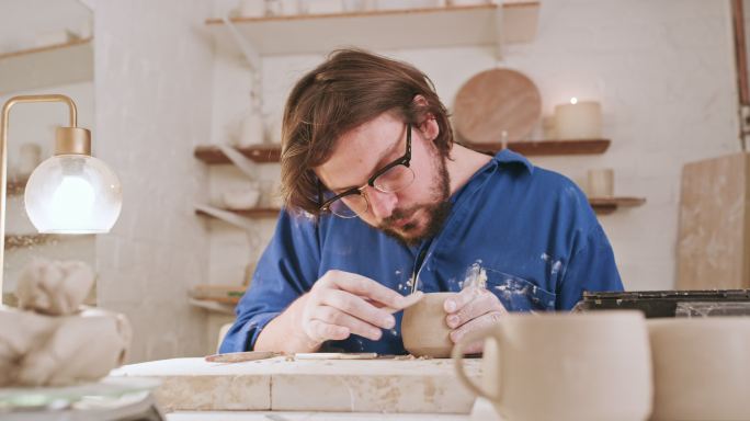 男雕塑家在他的陶器作坊里用工具雕刻一个陶罐。陶瓷艺术家、陶艺家或手工艺人，制作手工杯子或碗，为自己的