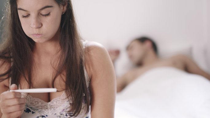 4k视频画面显示，一名年轻女子在接受怀孕测试后看起来很紧张，而她的丈夫在家里的背景下睡在床上